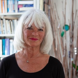 An older woman with white hair and a black top stands indoors in front of a bookshelf and decorative branches, smiling at the camera.