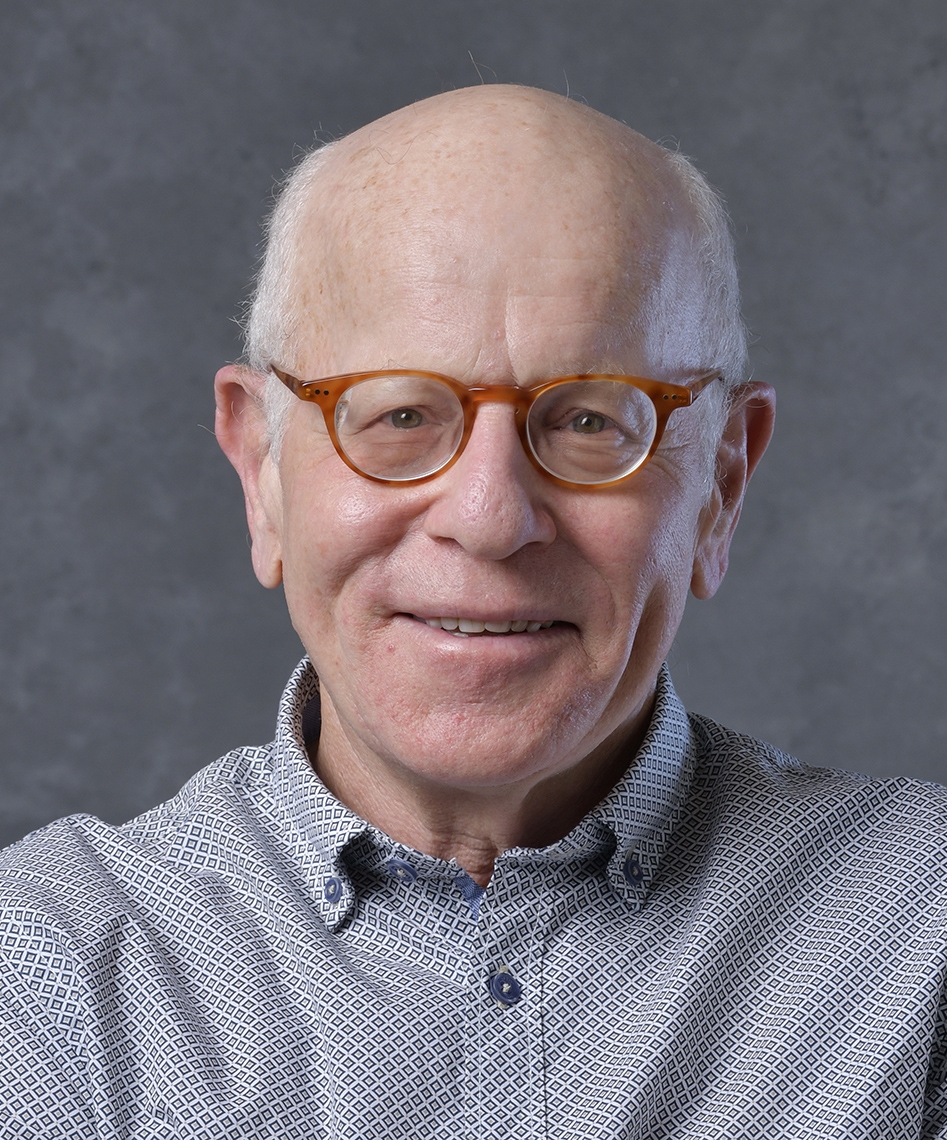 An older man with glasses and a patterned shirt smiles at the camera against a gray background.