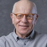 An older man with glasses and a patterned shirt smiles at the camera against a gray background.