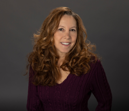 Woman with long, curly brown hair wearing a purple sweater, smiling at the camera against a plain dark background.
