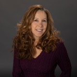 Woman with long, curly brown hair wearing a purple sweater, smiling at the camera against a plain dark background.