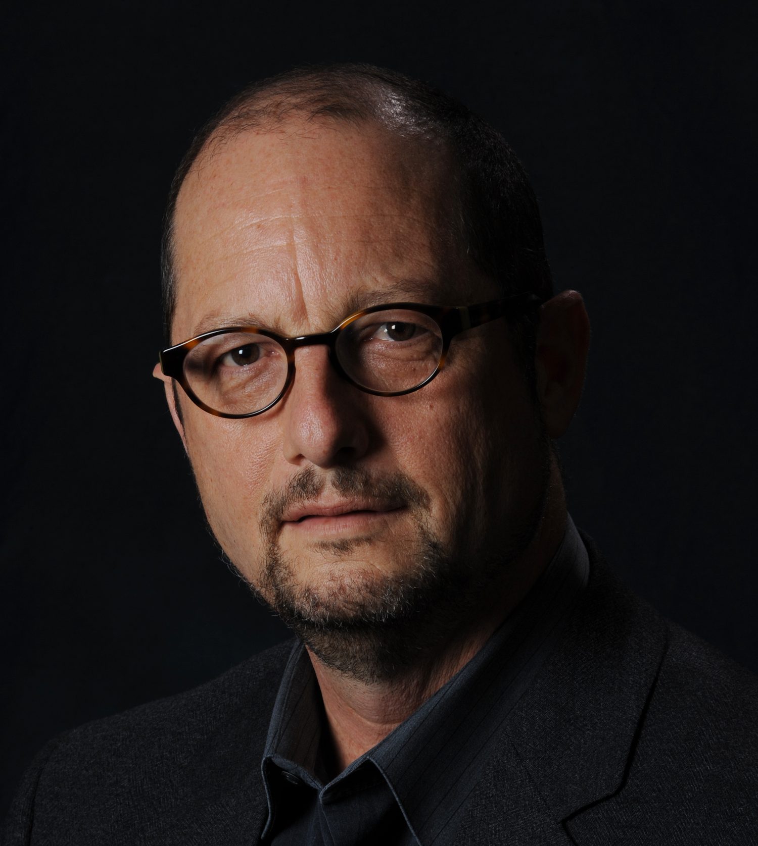 A middle-aged man with glasses, short dark hair, and a trimmed beard, wearing a dark blazer and shirt, poses against a dark background.