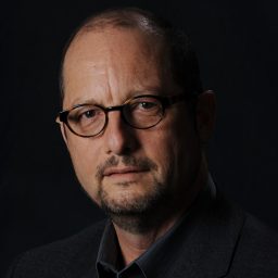 A middle-aged man with glasses, short dark hair, and a trimmed beard, wearing a dark blazer and shirt, poses against a dark background.