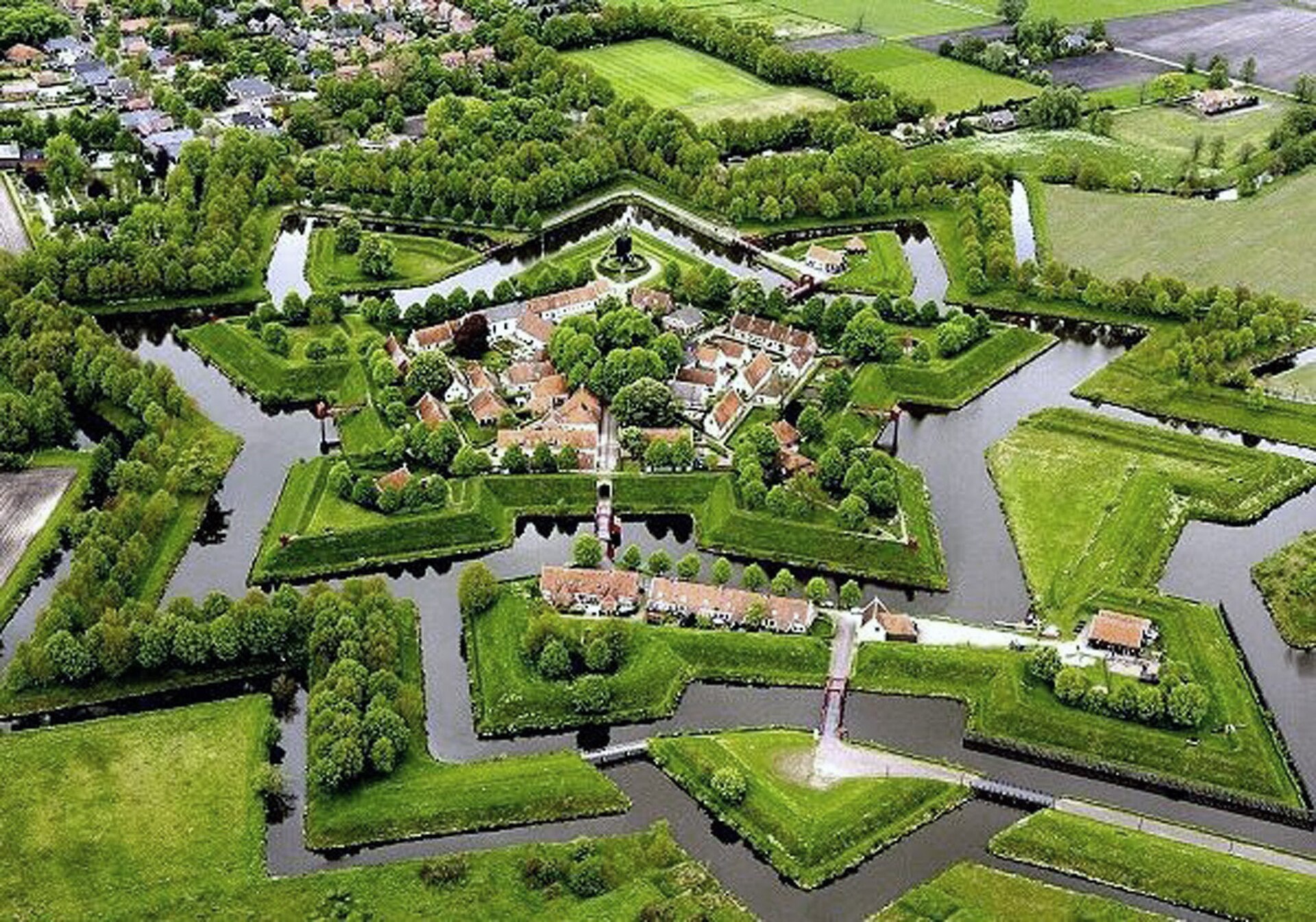 Aerial view of a star-shaped fort with red-roofed buildings, green lawns, trees, and surrounding moats, set in a rural landscape.