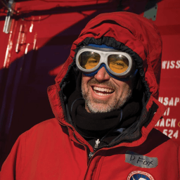 A person in a red parka and yellow-tinted goggles smiles outdoors in a cold environment; a name tag on the coat reads "D. Fox.