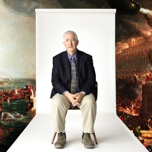 An older man in a suit sits on a chair in front of a backdrop showing a dramatic classical painting of chaos and destruction.