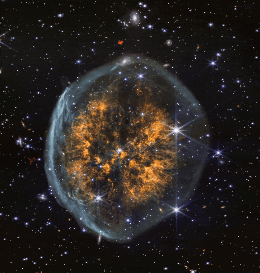 Colorful nebula with an orange, web-like center and a blue outer shell, surrounded by stars and galaxies against a black space background.