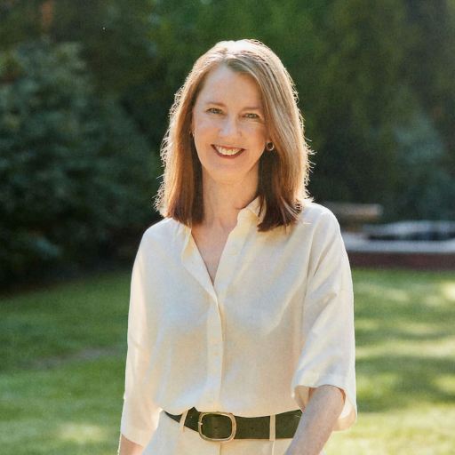 A woman with shoulder-length hair, wearing a white shirt and black belt, stands outdoors on a sunny day with grass and trees in the background.