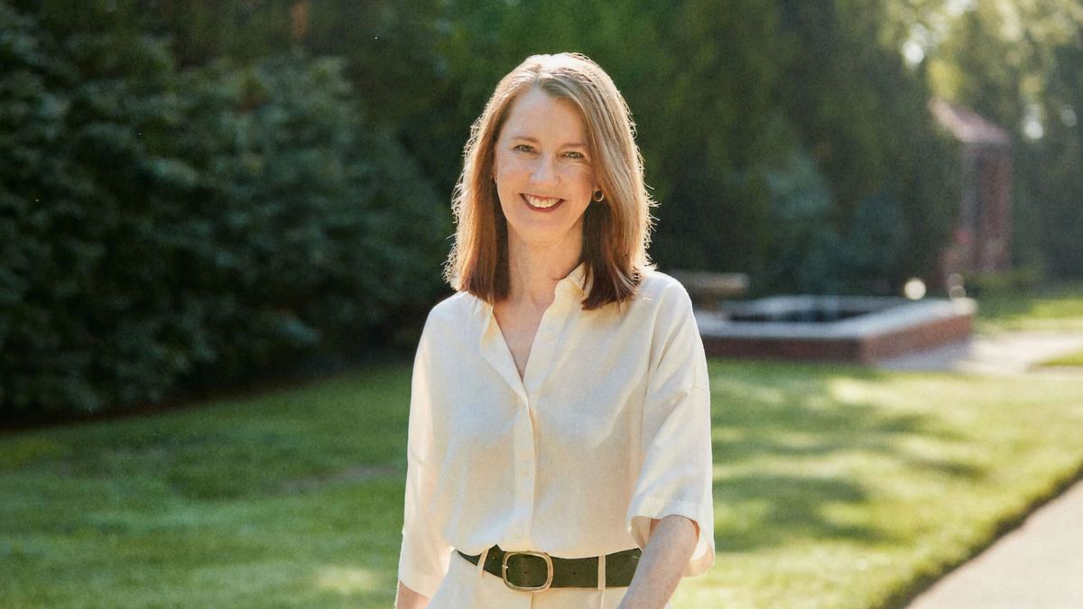A woman with shoulder-length hair, wearing a white shirt and black belt, stands outdoors on a sunny day with grass and trees in the background.