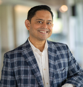 A man wearing a blue and white plaid blazer and white shirt smiles while standing indoors in a well-lit hallway.