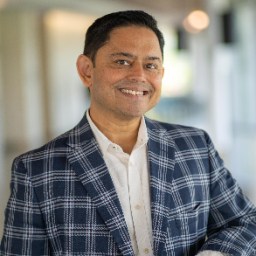 A man wearing a blue and white plaid blazer and white shirt smiles while standing indoors in a well-lit hallway.