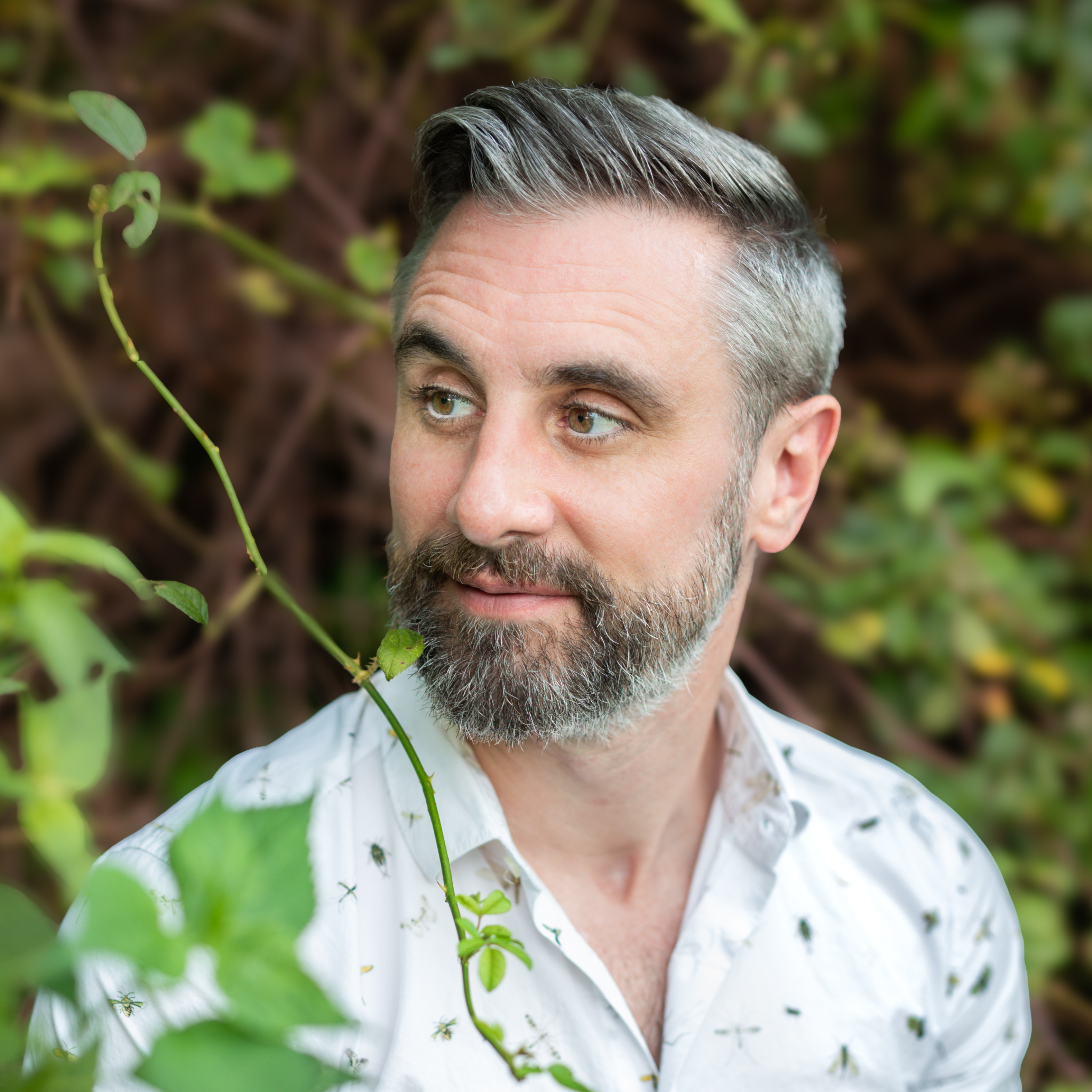 A man with gray hair and a beard wearing a white patterned shirt stands outdoors, looking to the side with greenery in the background.