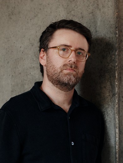 A man with short brown hair, a beard, and glasses, wearing a dark shirt, stands against a concrete wall.
