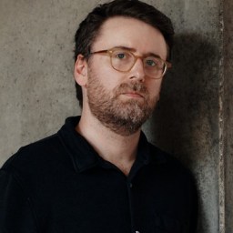 A man with short brown hair, a beard, and glasses, wearing a dark shirt, stands against a concrete wall.
