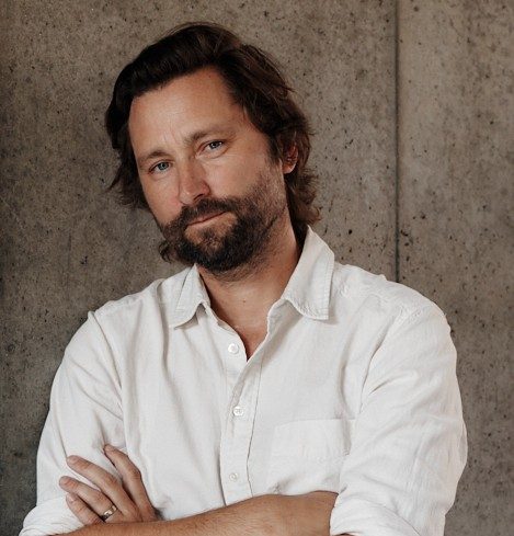 A man with wavy brown hair and a beard stands against a concrete wall, wearing a white button-up shirt with his arms crossed.