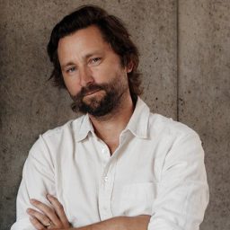 A man with wavy brown hair and a beard stands against a concrete wall, wearing a white button-up shirt with his arms crossed.