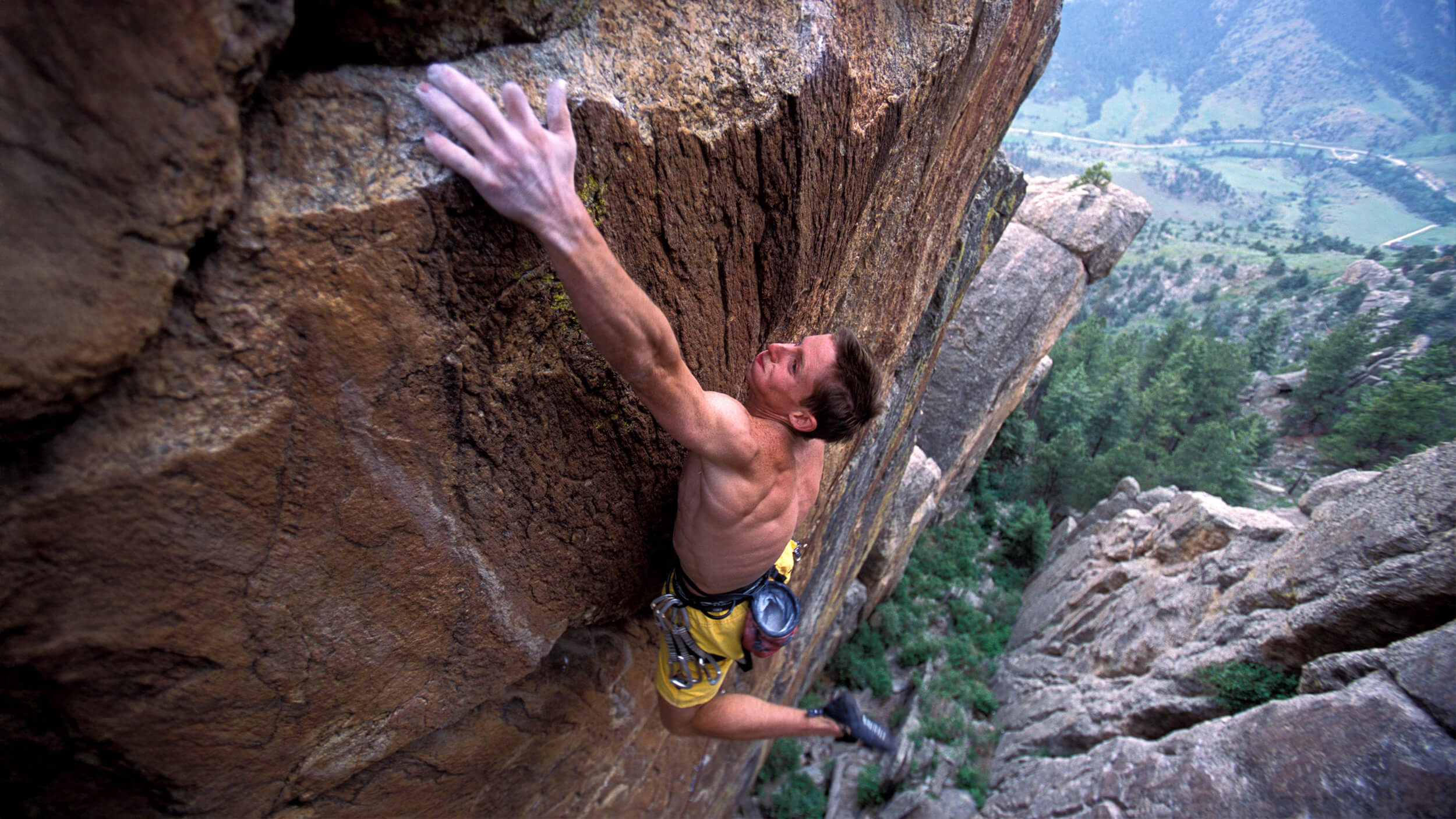 A shirtless man, resembling Tommy Caldwell, climbs a steep rock face high above the ground, reaching for a hold with one hand and gripping the rock with the other; trees and a valley stretch out below.