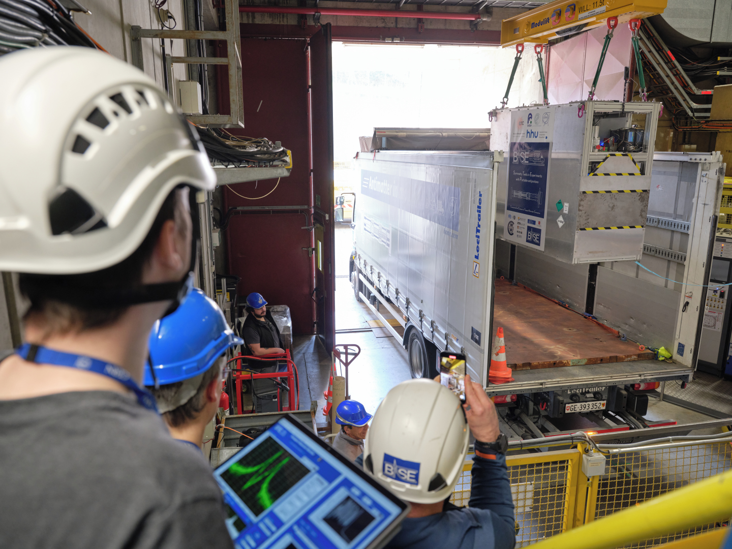 Workers wearing helmets supervise the loading of a large container&mdash;housing equipment for antimatter interstellar travel&mdash;into a truck using lifting gear inside an industrial facility.