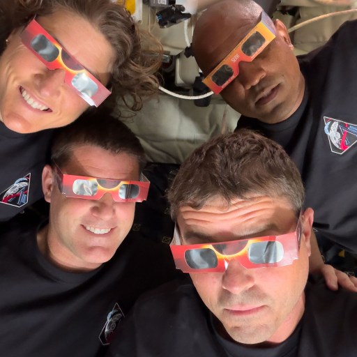 Four people wearing black shirts and eclipse glasses look up at the camera indoors, their excitement echoing the spirit of the Artemis II distance record mission.