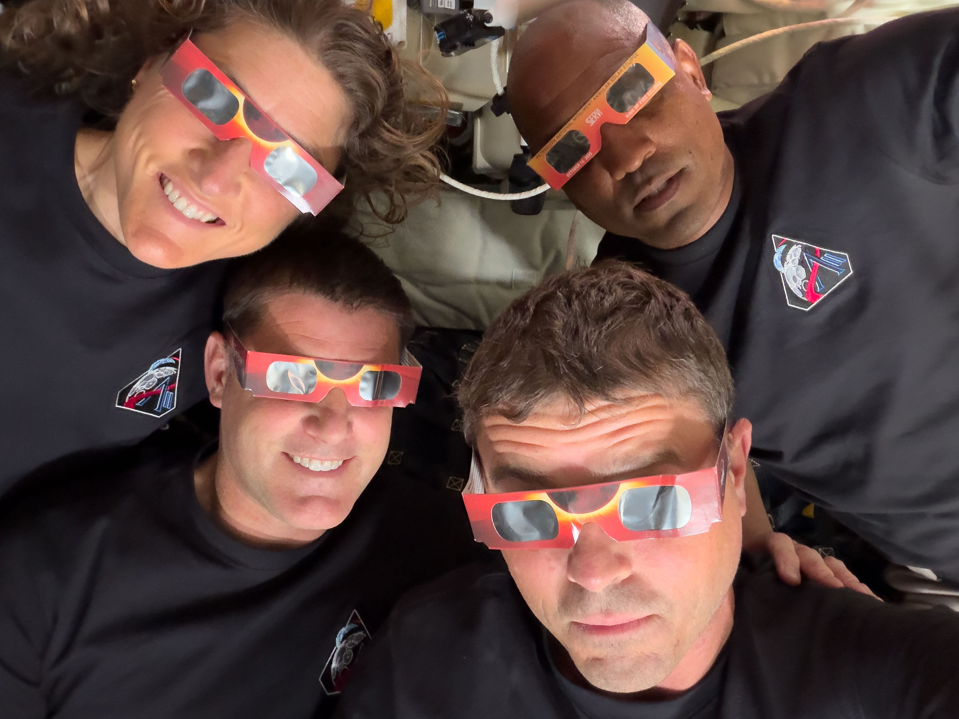 Four people wearing black shirts and eclipse glasses look up at the camera indoors, their excitement echoing the spirit of the Artemis II distance record mission.