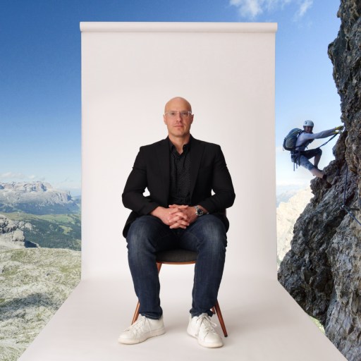 A man in casual business attire sits on a chair in front of a white backdrop, with a mountain landscape and a climber scaling a rocky cliff in the background.