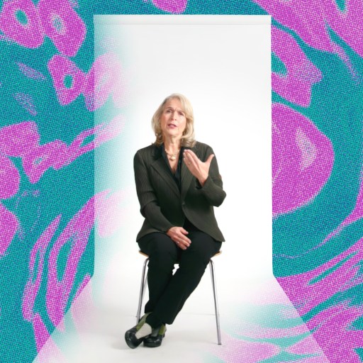 A woman in a black suit sits on a stool in a white room, gesturing with one hand. The background features abstract teal and pink patterns.