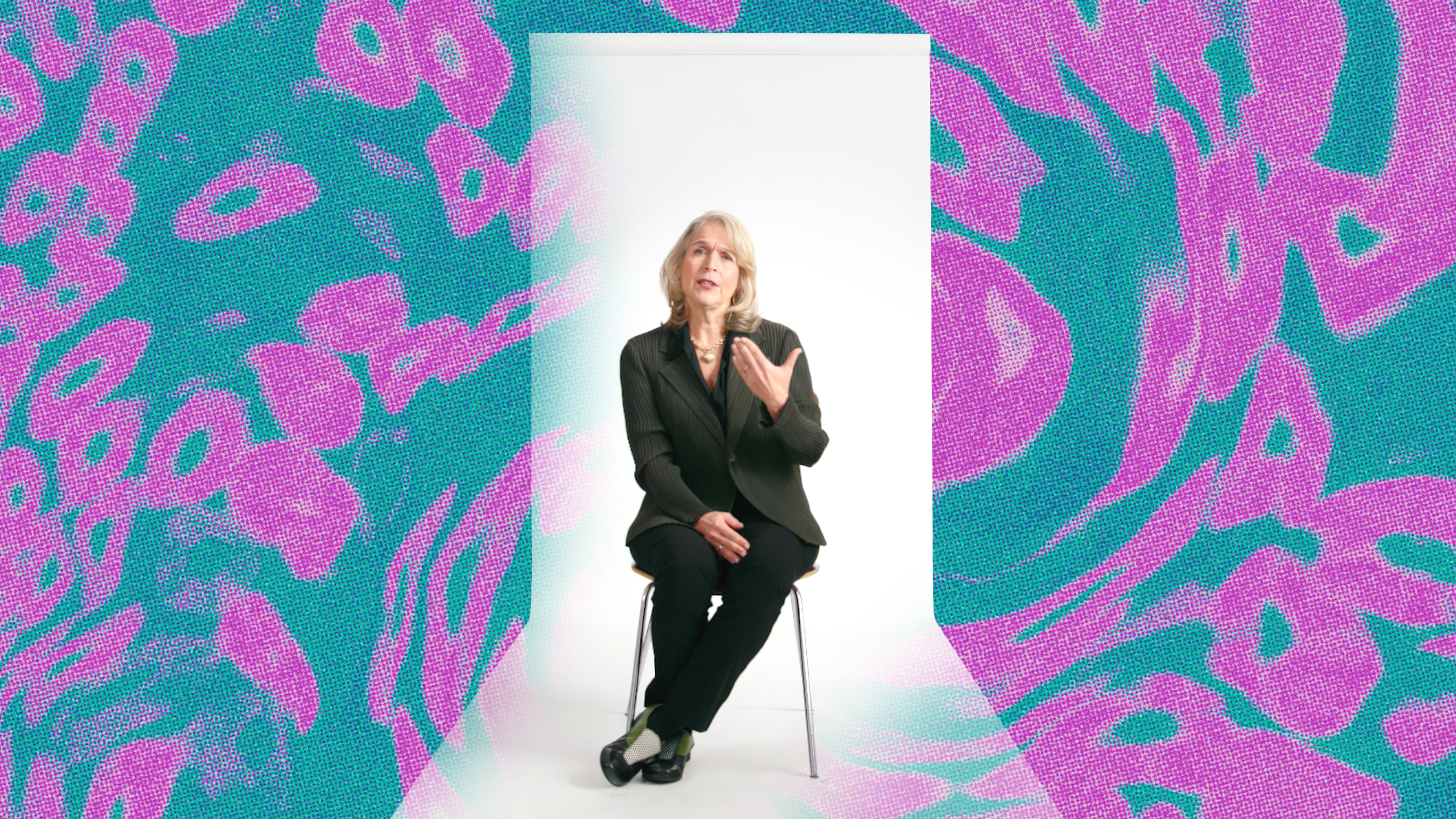 A woman in a black suit sits on a stool in a white room, gesturing with one hand. The background features abstract teal and pink patterns.