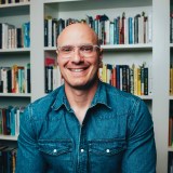 A person wearing glasses and a denim shirt smiles while sitting in front of bookshelves filled with books.