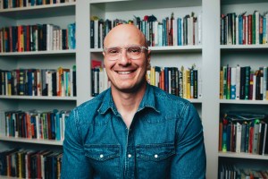 A person wearing glasses and a denim shirt smiles while sitting in front of bookshelves filled with books.