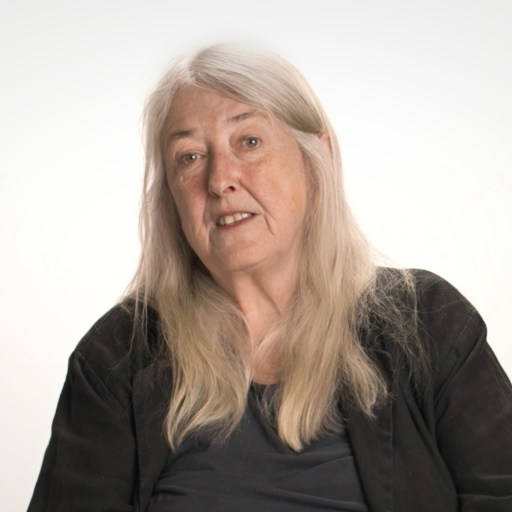An older woman with long gray hair wearing a dark jacket and shirt sits against a plain, light background, looking slightly toward the camera.