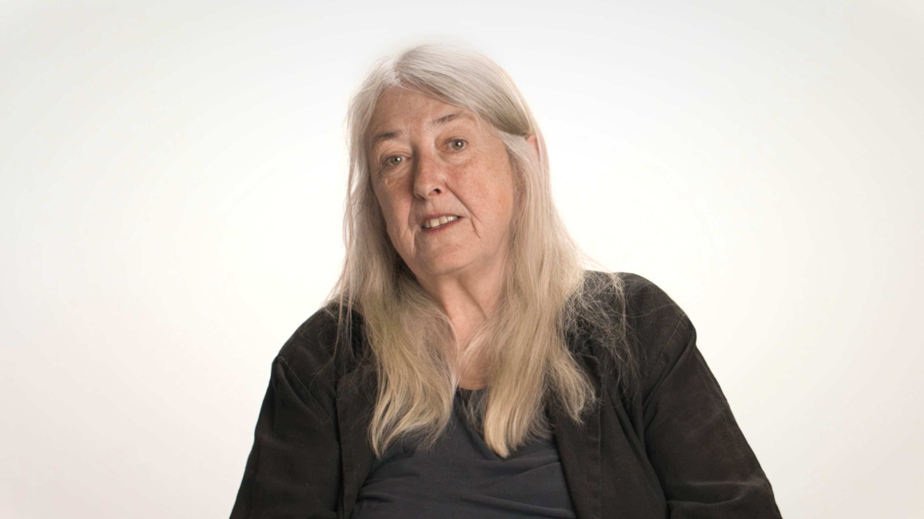 An older woman with long gray hair wearing a dark jacket and shirt sits against a plain, light background, looking slightly toward the camera.
