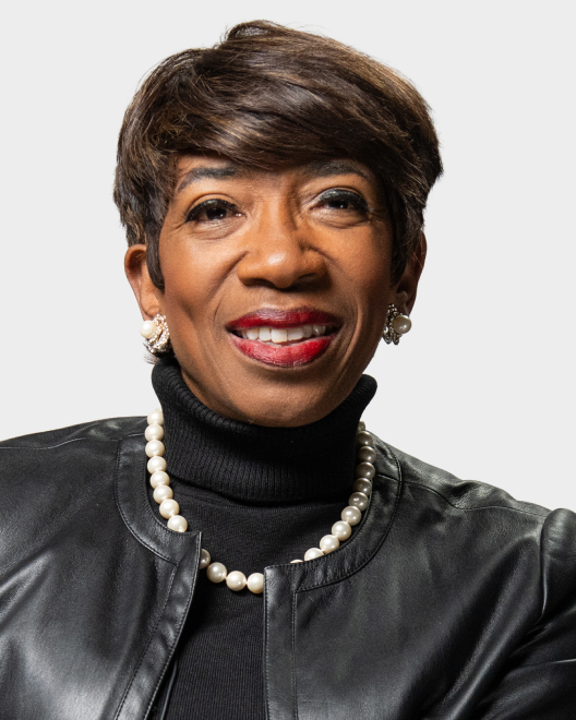 A woman with short brown hair, wearing a black leather jacket, black turtleneck, pearl necklace, pearl earrings, and red lipstick, smiles in front of a plain light background.