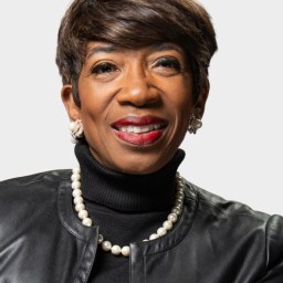 A woman with short brown hair, wearing a black leather jacket, black turtleneck, pearl necklace, pearl earrings, and red lipstick, smiles in front of a plain light background.