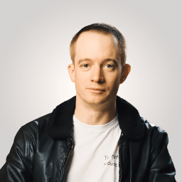 A man with short hair wearing a black leather jacket over a white t-shirt, sitting against a plain light gray background.