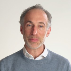 A middle-aged man with short gray hair, light skin, and a trimmed beard wearing a light blue sweater over a white collared shirt, standing against a plain light background.