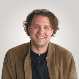 Man with wavy brown hair wearing a brown jacket over a black shirt, smiling in front of a plain light background.