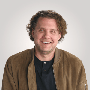 Man with wavy brown hair wearing a brown jacket over a black shirt, smiling in front of a plain light background.
