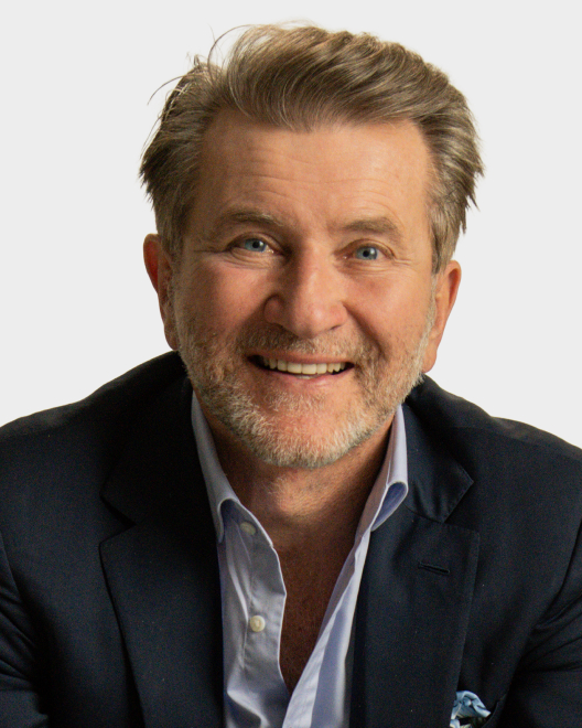 A man with light brown hair and a short beard wearing a dark suit jacket and light blue shirt, smiling at the camera against a plain background.