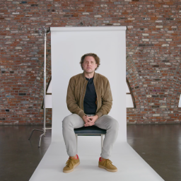 A man sits on a chair in front of a white backdrop in a brick-walled room with arched windows; "B T" logo is visible in the top right corner.