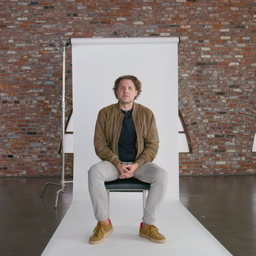A man sits on a chair in front of a white backdrop in a brick-walled room with arched windows; "B T" logo is visible in the top right corner.
