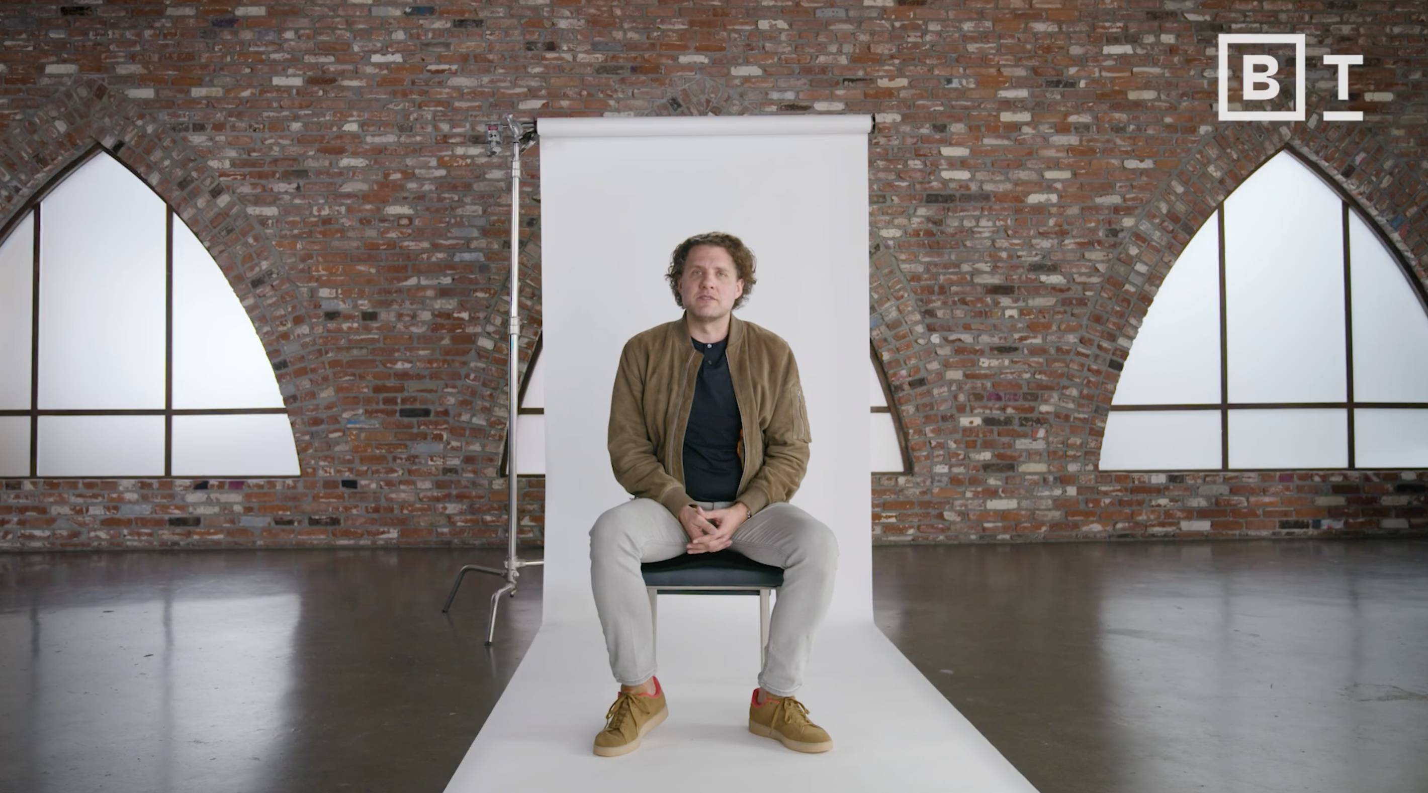 A man sits on a chair in front of a white backdrop in a brick-walled room with arched windows; "B T" logo is visible in the top right corner.