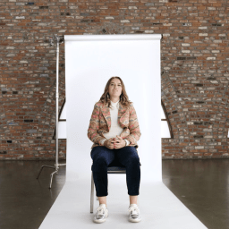 A woman sits on a chair in front of a white backdrop in a brick-walled room with arched windows. The BT logo is visible in the top right corner.