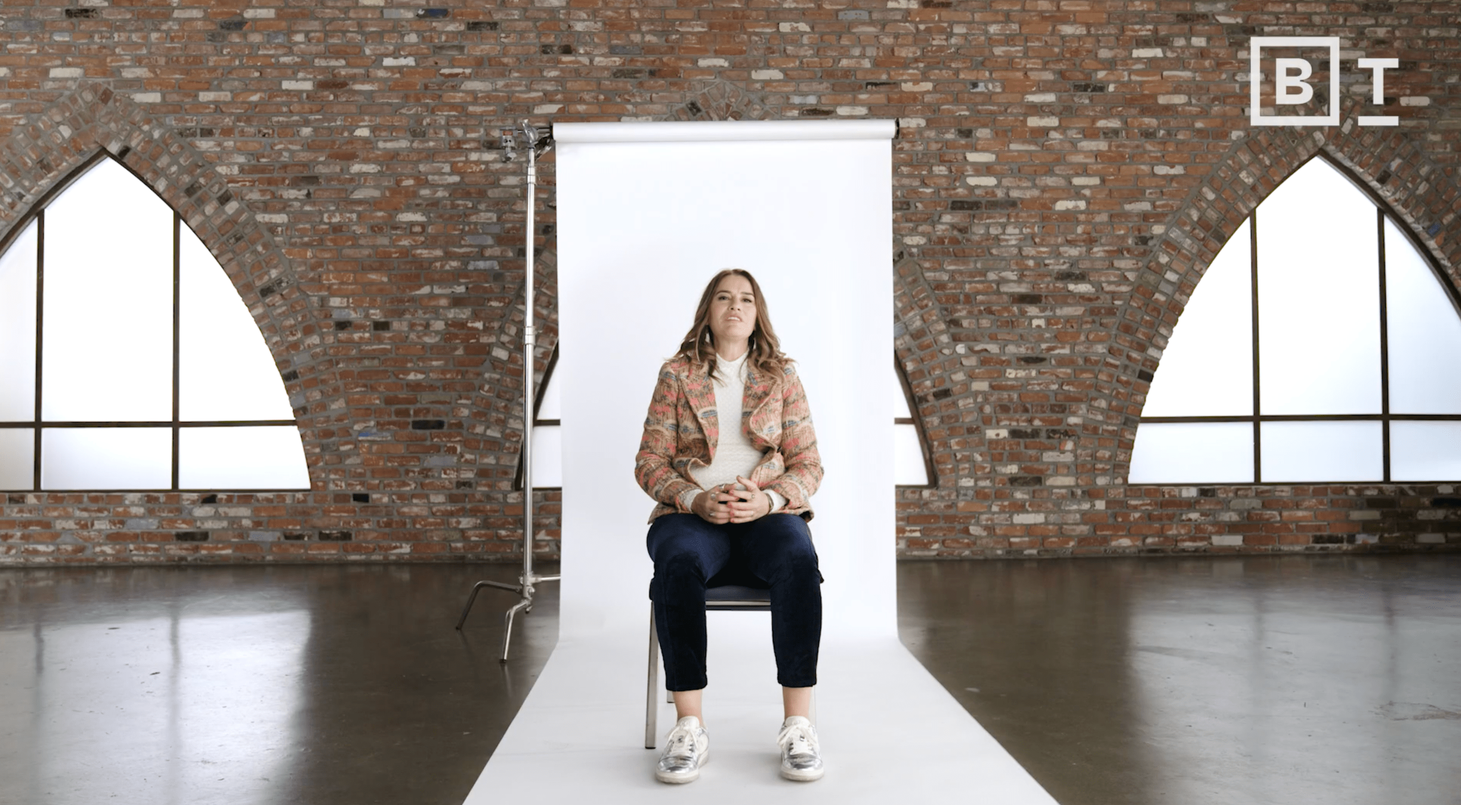A woman sits on a chair in front of a white backdrop in a brick-walled room with arched windows. The BT logo is visible in the top right corner.