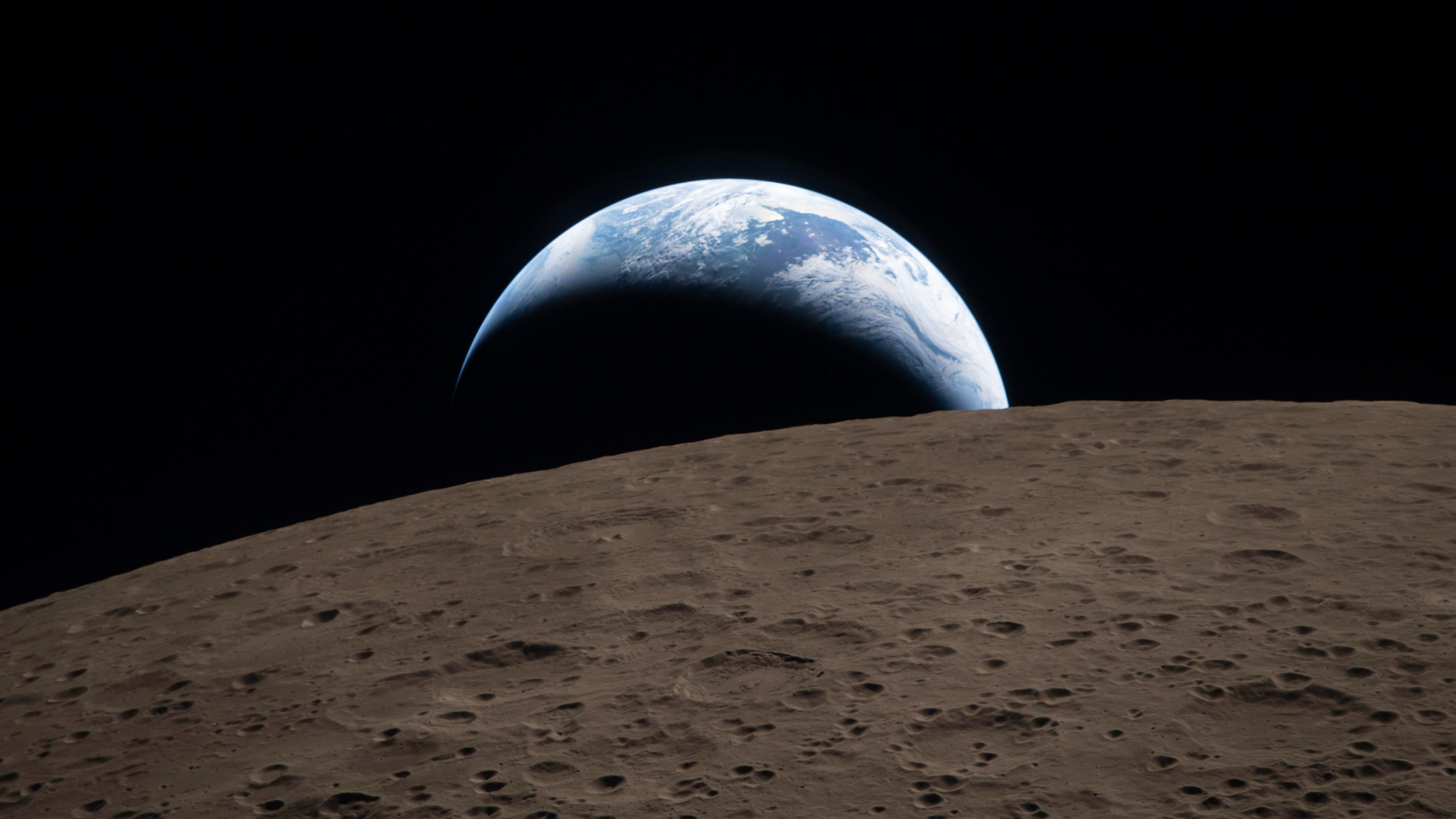 Earth rises above the horizon of the Moon, with the lunar surface in the foreground and space in the background.