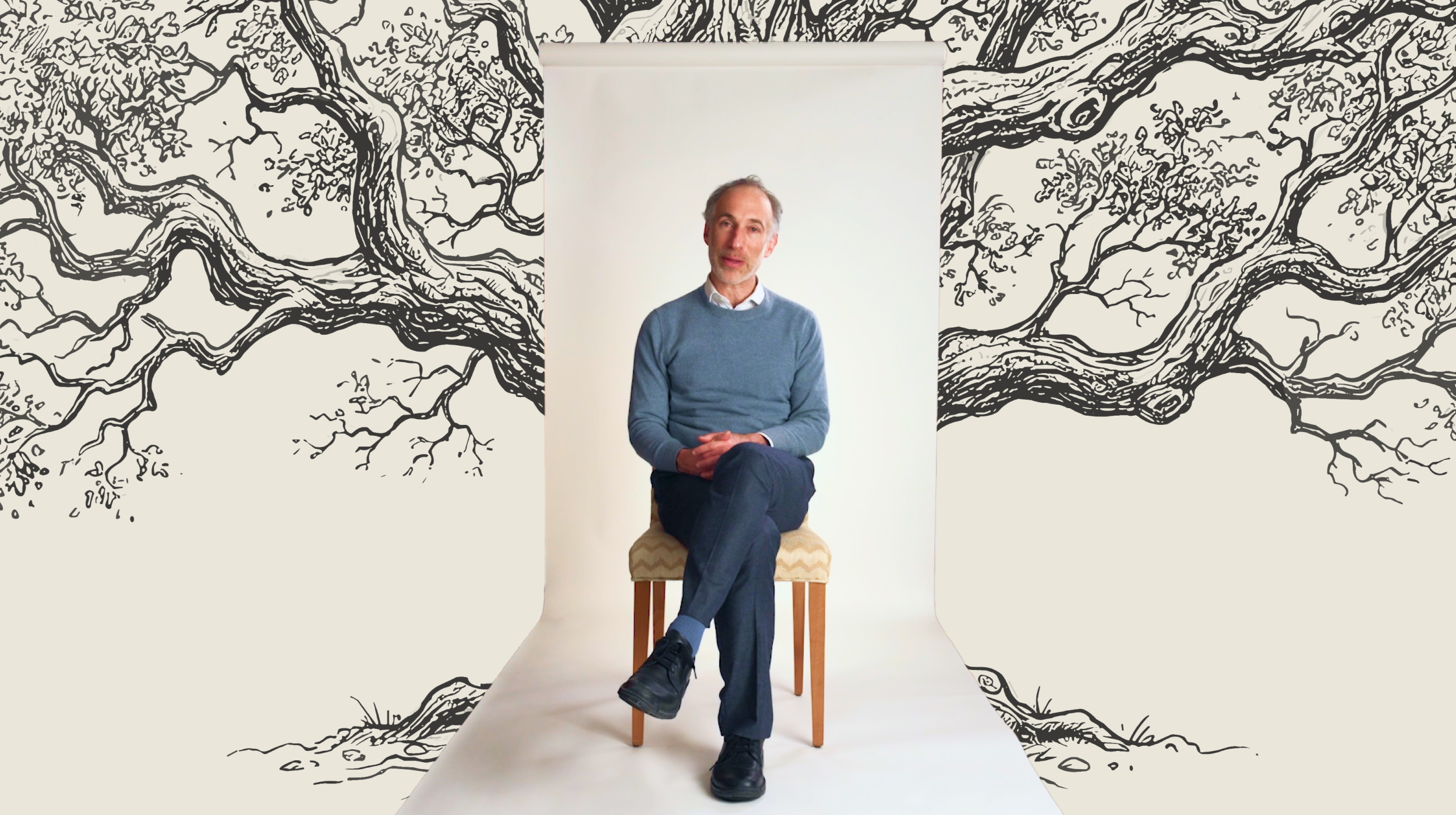 A man in a blue sweater and dark pants sits on a chair against a white backdrop with illustrated tree branches in the background.