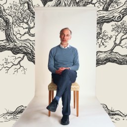 A man in a blue sweater and dark pants sits on a chair against a white backdrop with illustrated tree branches in the background.