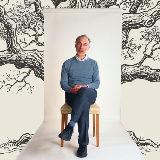 A man in a blue sweater and dark pants sits on a chair against a white backdrop with illustrated tree branches in the background.