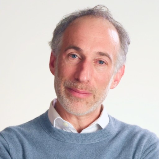 Middle-aged man with gray hair and beard, wearing a light blue sweater over a white shirt, standing against a plain light background, looking at the camera.
