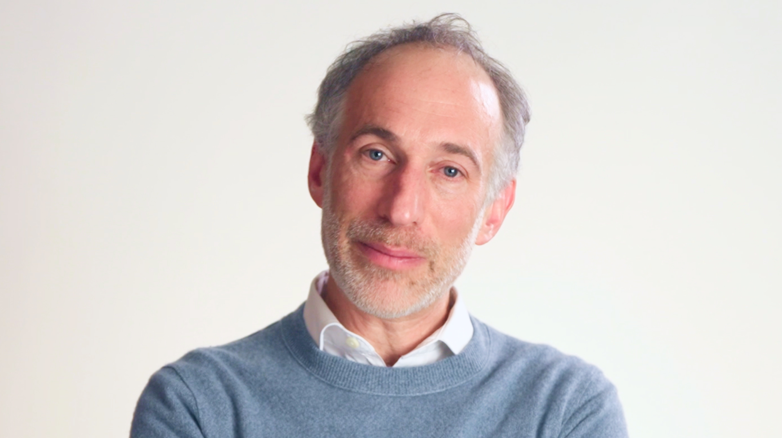 Middle-aged man with gray hair and beard, wearing a light blue sweater over a white shirt, standing against a plain light background, looking at the camera.