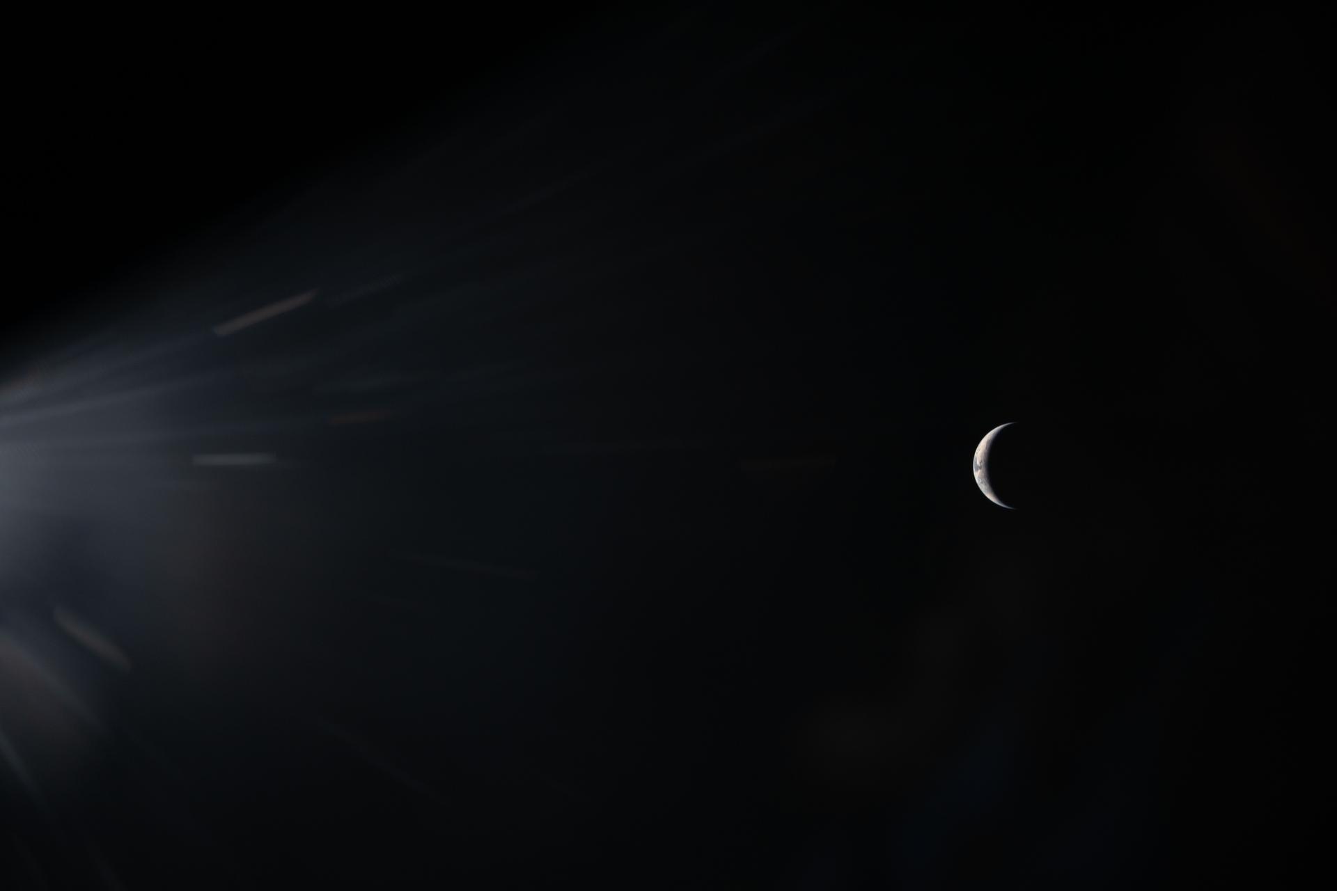 A crescent moon appears on the right side of a dark sky, with light rays streaming in from the left&mdash;echoing the hope and wonder inspired by Artemis II as it journeys between Earth and the Moon.